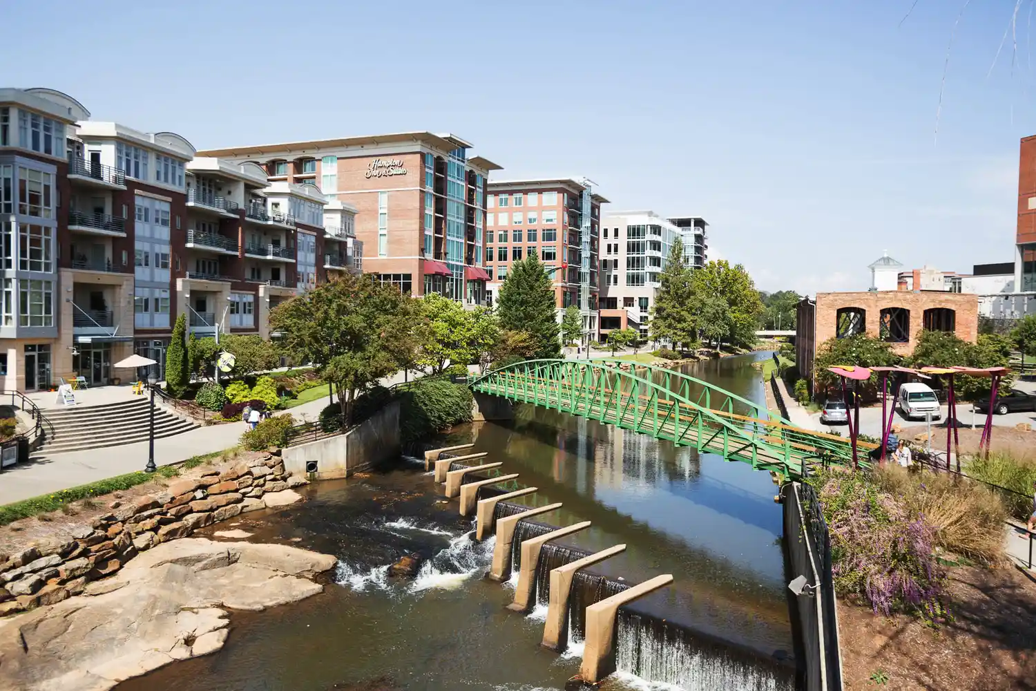 A modern pedestrian bridge crossing a river in a lush green urban park in downtown Greenville.