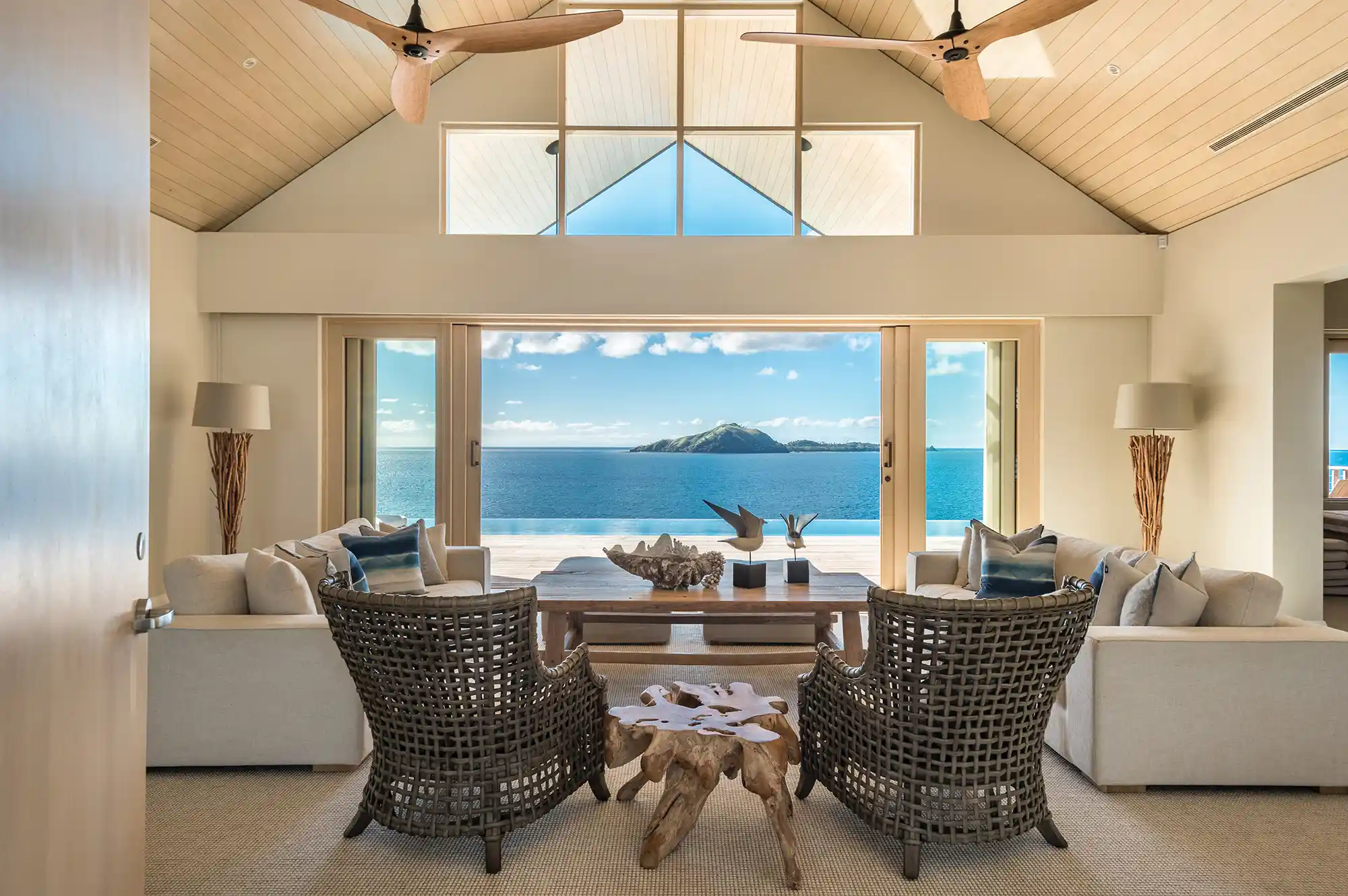 Luxurious living space with wicker chairs and tropical decor facing wide-open windows with an ocean view.