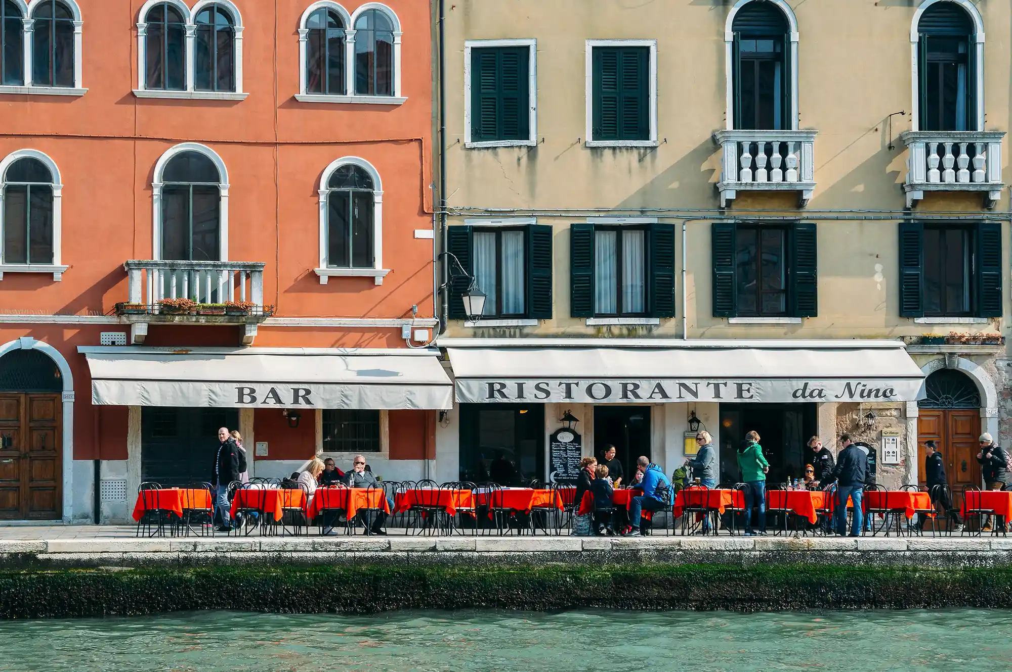Outdoor terrace of a Venetian restaurant along a canal with patrons dining during the off-season.