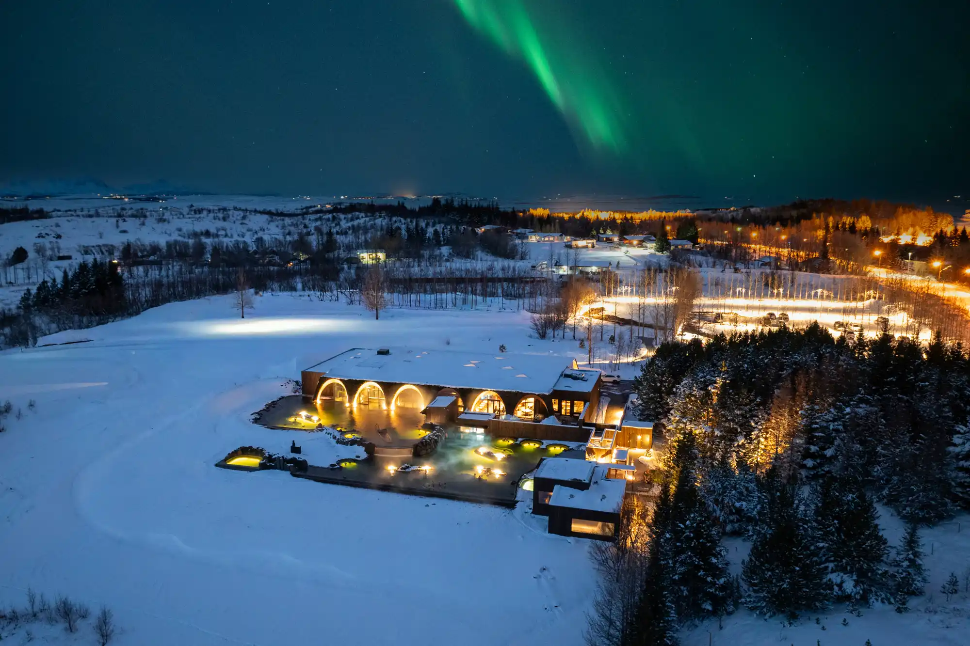 Aerial view of Laugarás Lagoon at night under the northern lights with snow-covered surroundings.