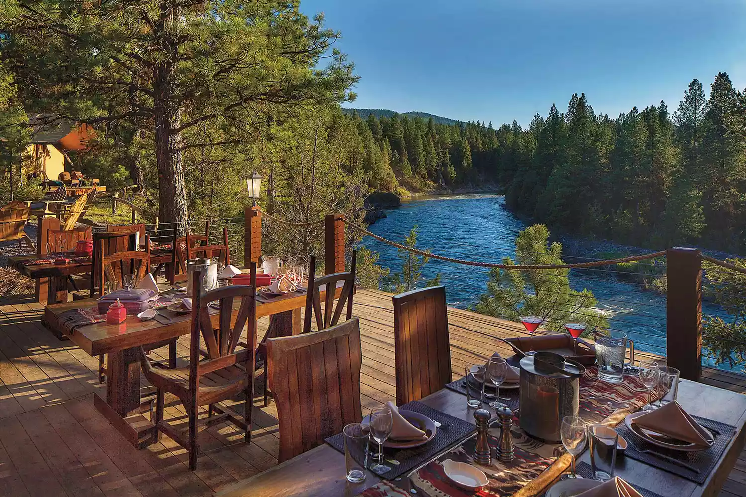 Outdoor dining area at a luxury riverside camp with white tablecloths and rustic scenery.