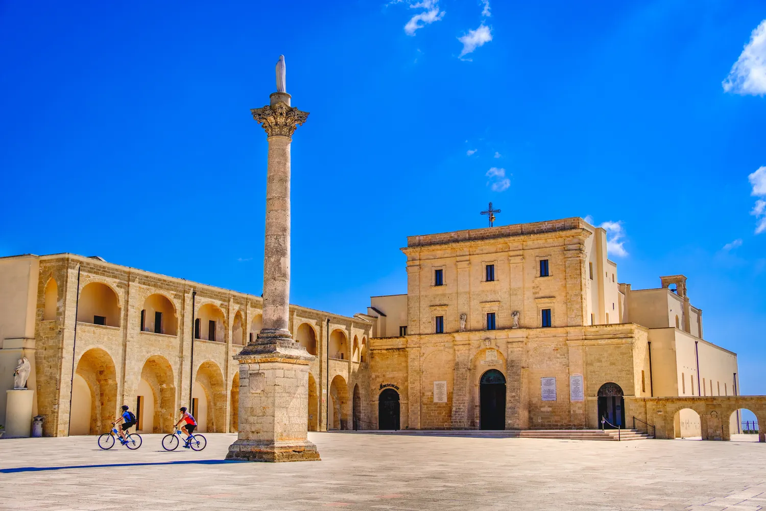 Two people cycling through a historic plaza with ancient landmarks in Lecce.