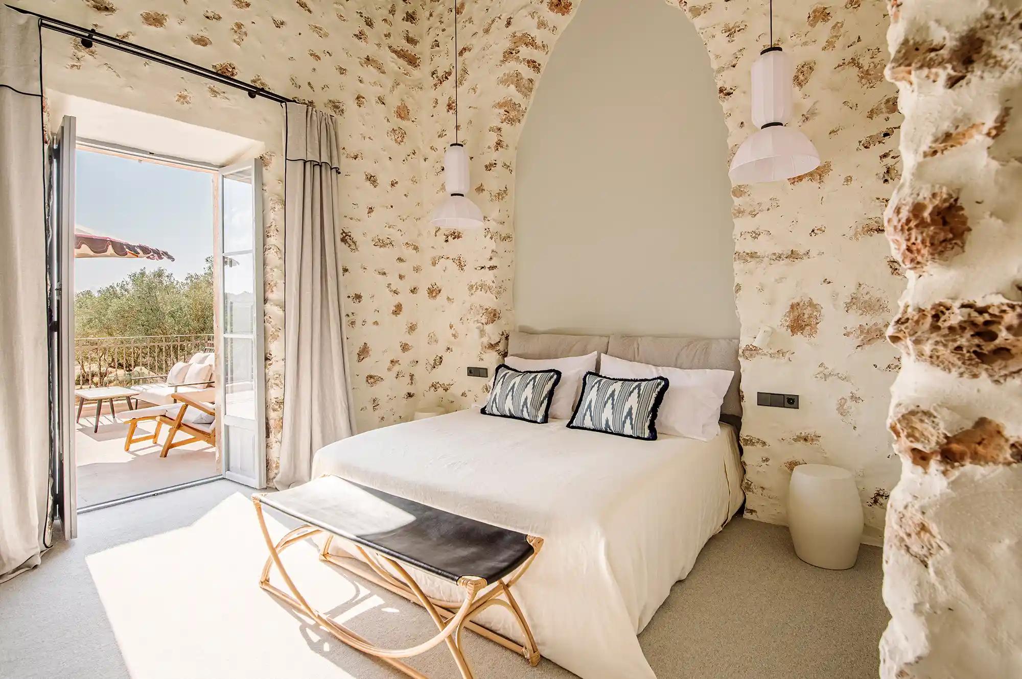 A master bedroom with an exposed stone wall and a view of a sunlit terrace through an open door.