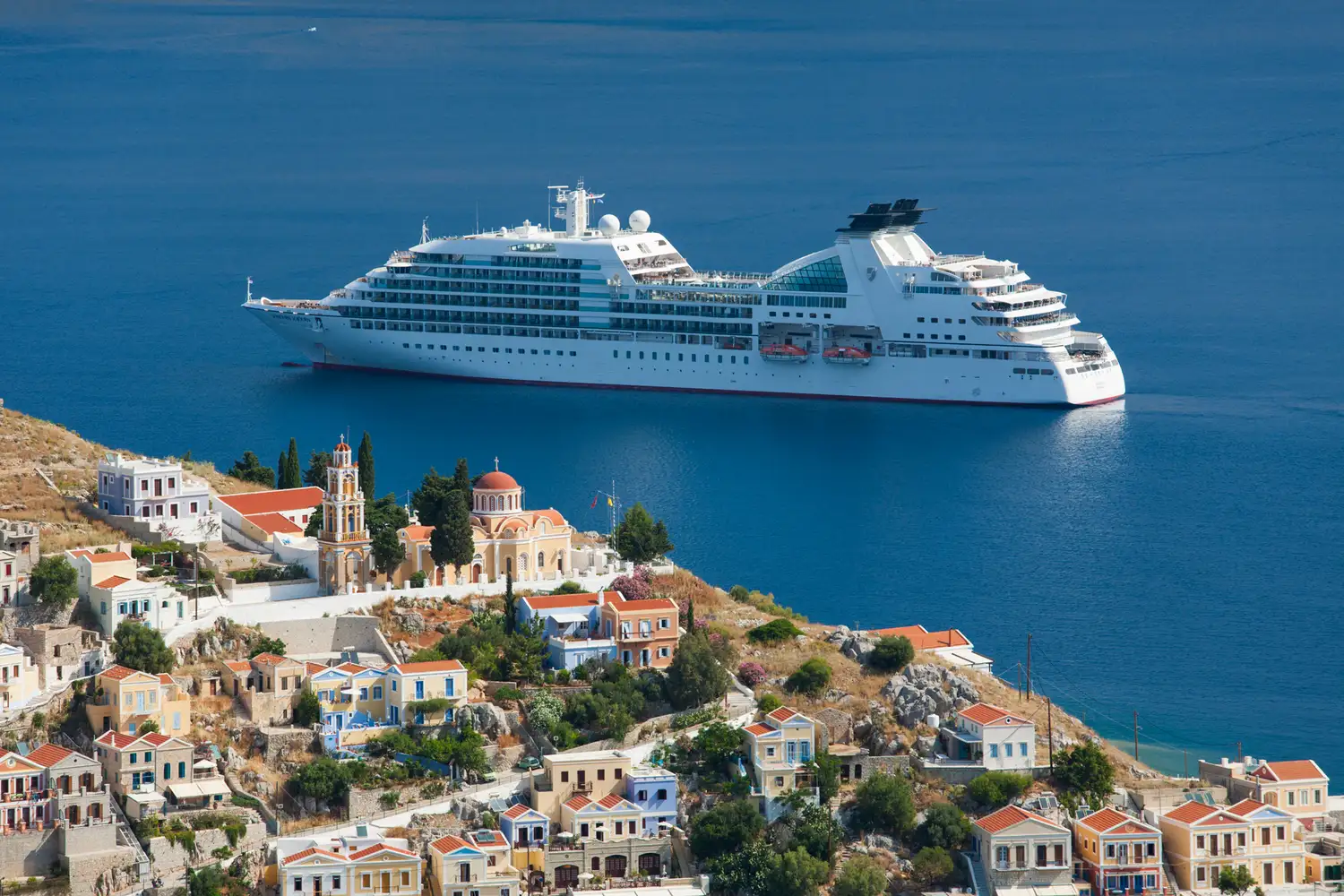 A large cruise ship docked near a scenic coastal town with traditional architecture and a church steeple visible