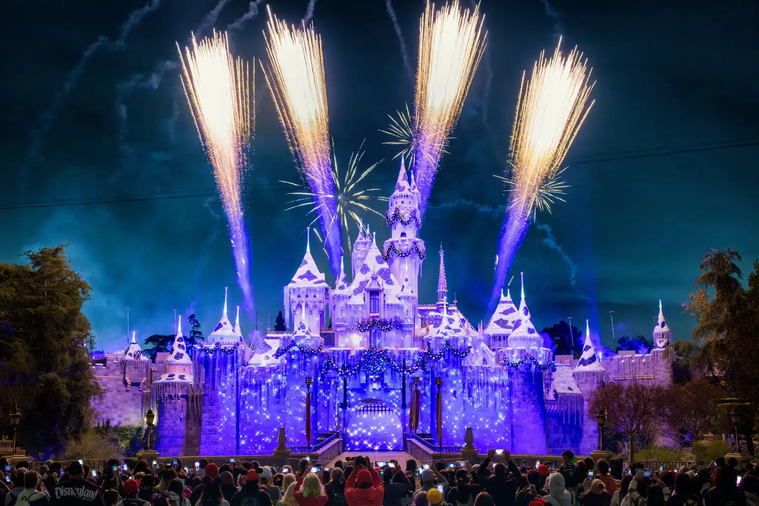 Fireworks exploding over a Disneyland castle during a nighttime holiday event with crowds watching.