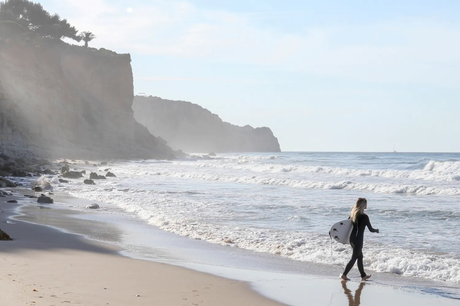 A person in a wetsuit carrying a surfboard toward the ocean with cliffs in the distance.