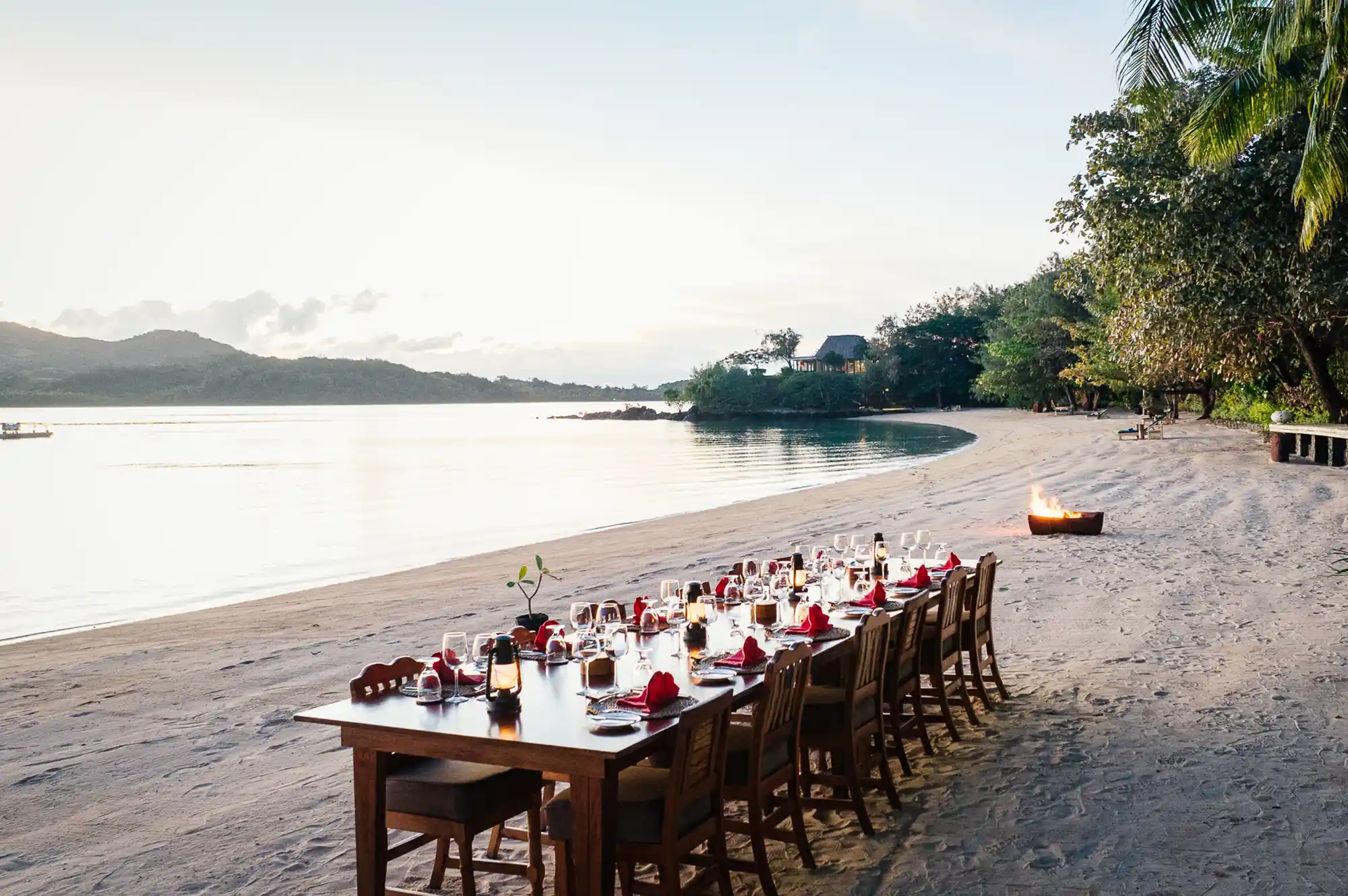 Dining table set for a group dinner on the beach at night with a fire pit nearby.