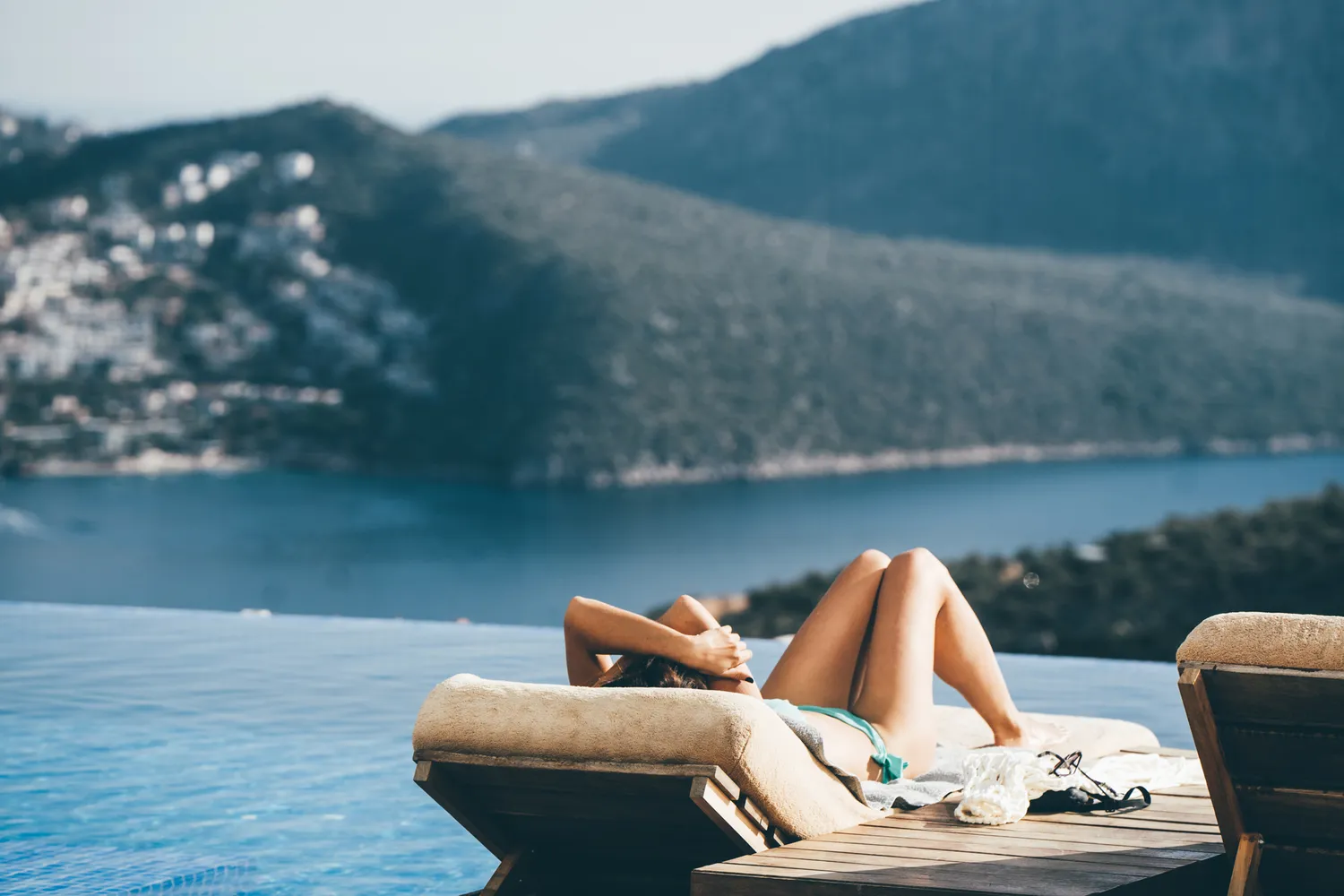 Person relaxing on a sun lounger beside an infinity pool overlooking a coastal view and distant hills