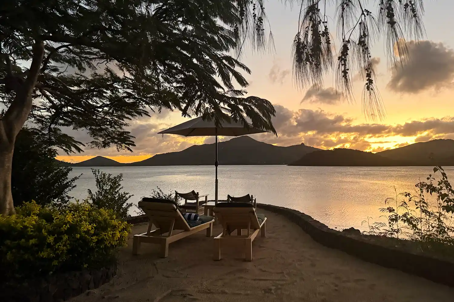 Two lounge chairs under a sunset sky on the porch of a thatched-roof bure.
