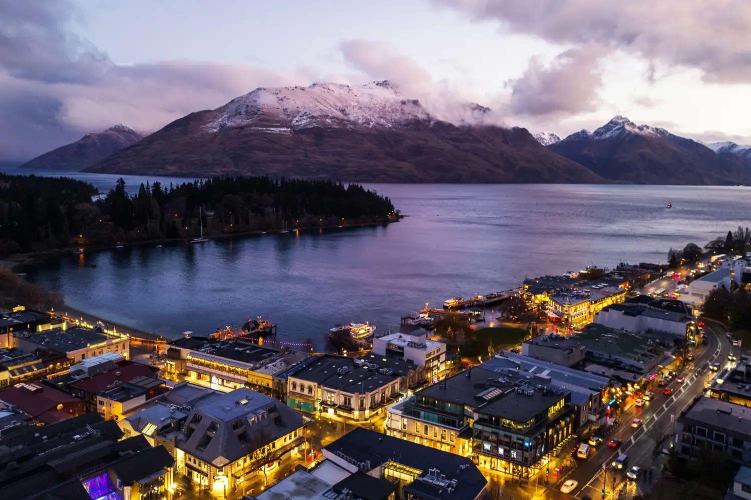 A twilight view of Queenstown's city lights nestled against the dark silhouettes of the Southern Alps and Lake Wakatipu.