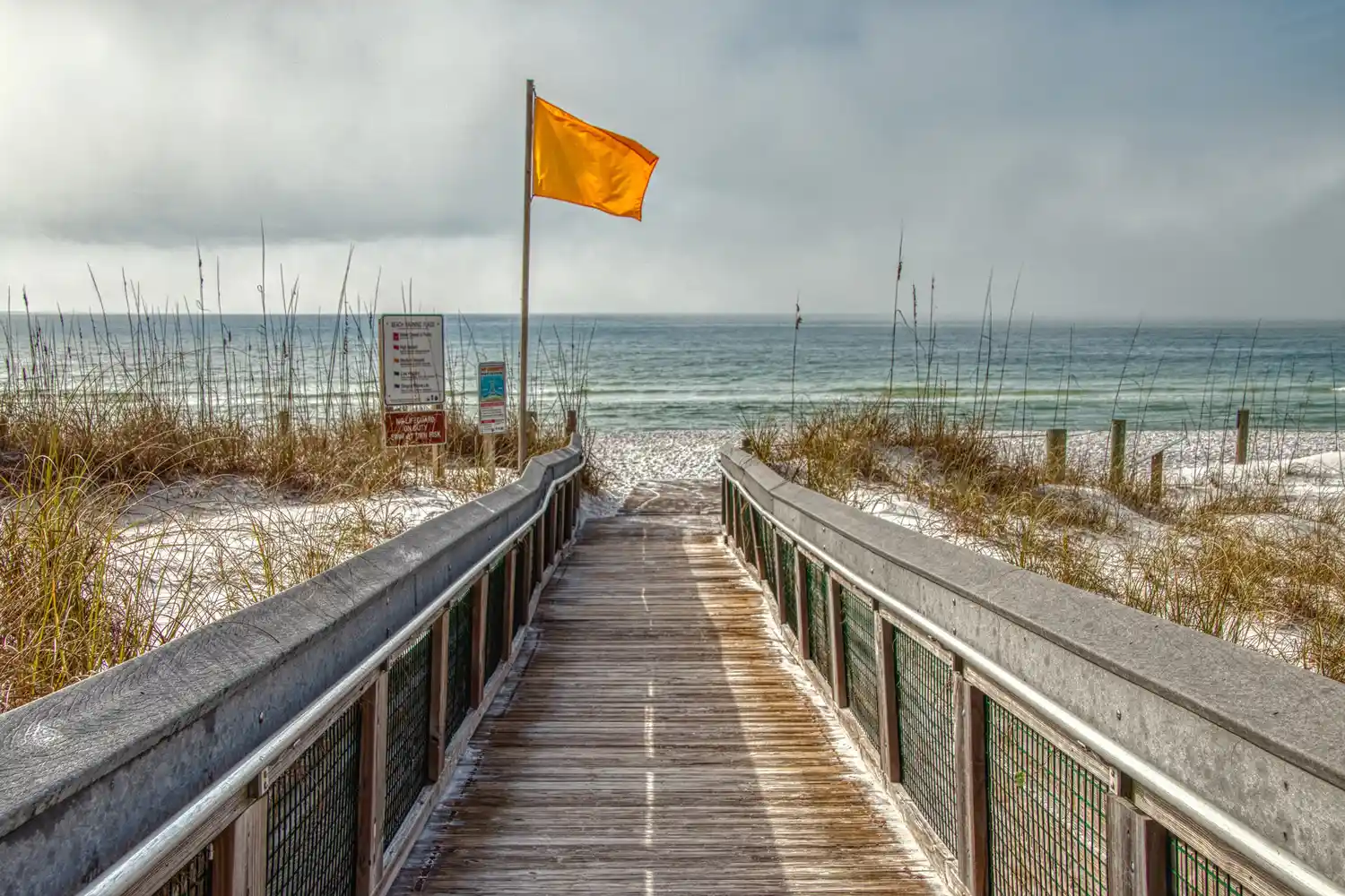 A wooden walkway leads to the beach entrance with a yellow safety flag flying.