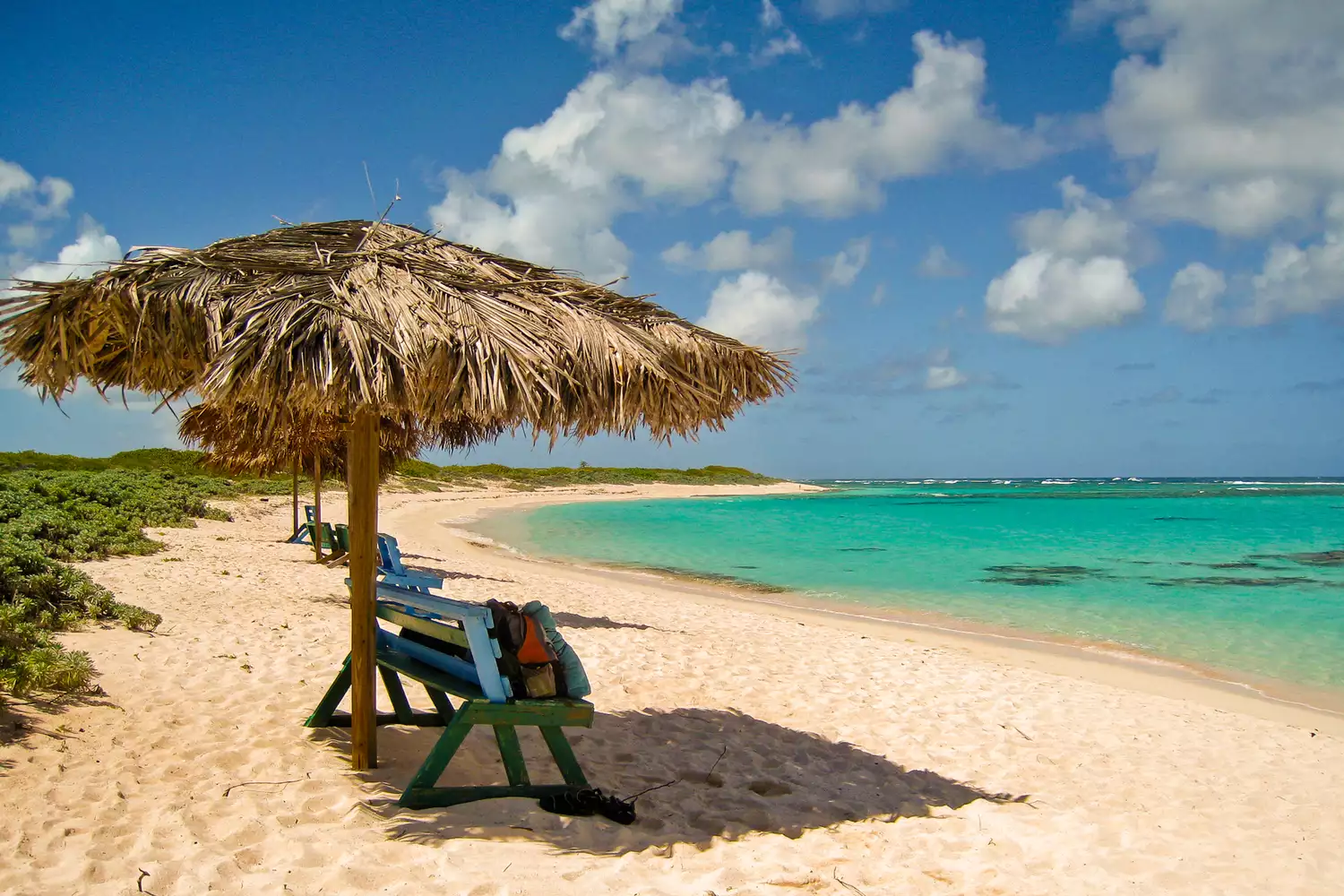 Thatched roof palapas with benches on a sandy beach overlook the crystal blue sea.