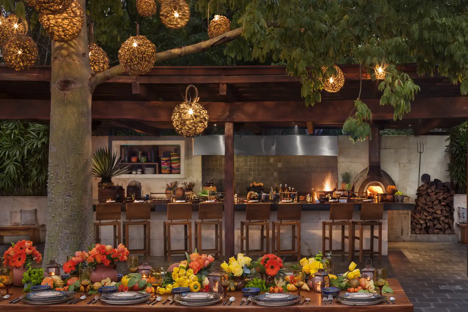 A long wooden communal dining table decorated with flowers in an outdoor garden kitchen setting.