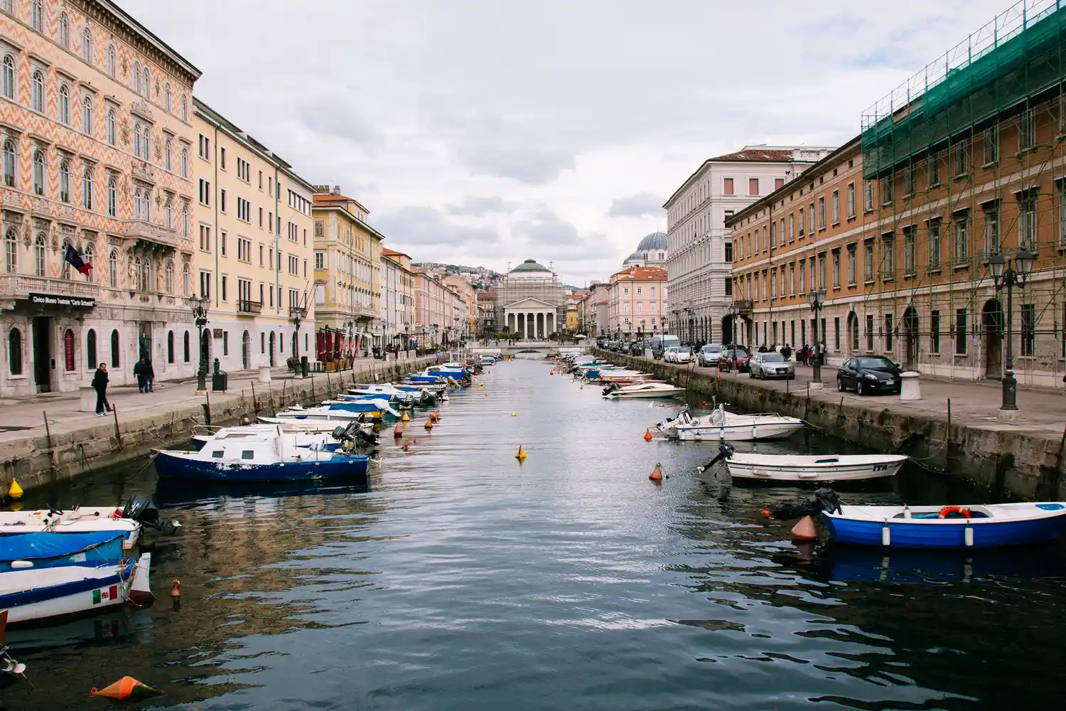 A peaceful canal in Trieste with moored boats and historic European buildings lining the water.