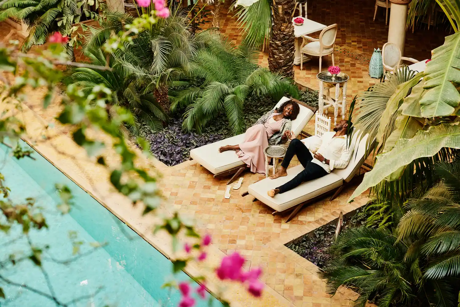 Two people relaxing on lounge chairs by a pool in a lush tropical setting surrounded by plants and flowers