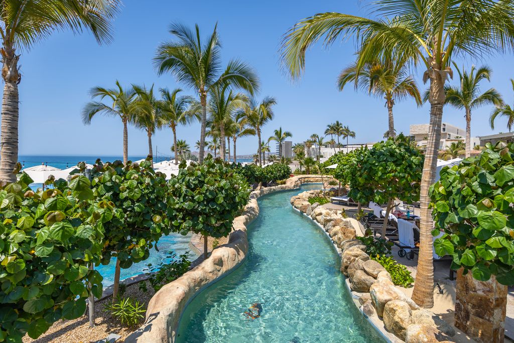Infinity pool and lazy river area at a beach resort with white umbrellas overlooking the blue sea.