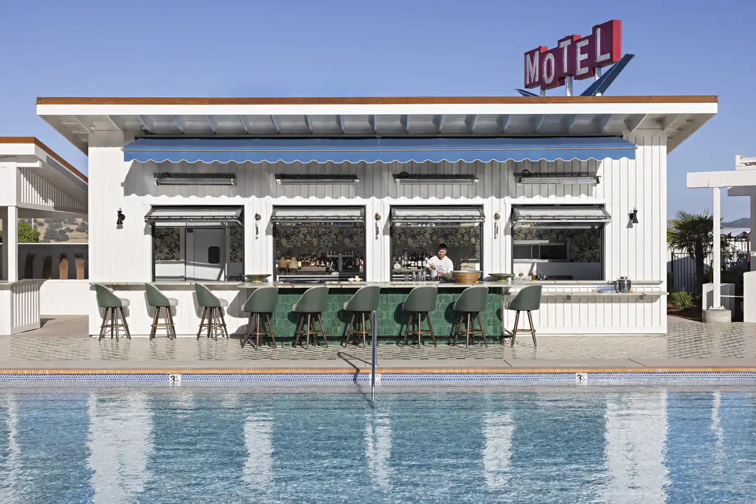 A bartender preparing a cocktail at a modern poolside bar with a vintage-style motel building in the background.