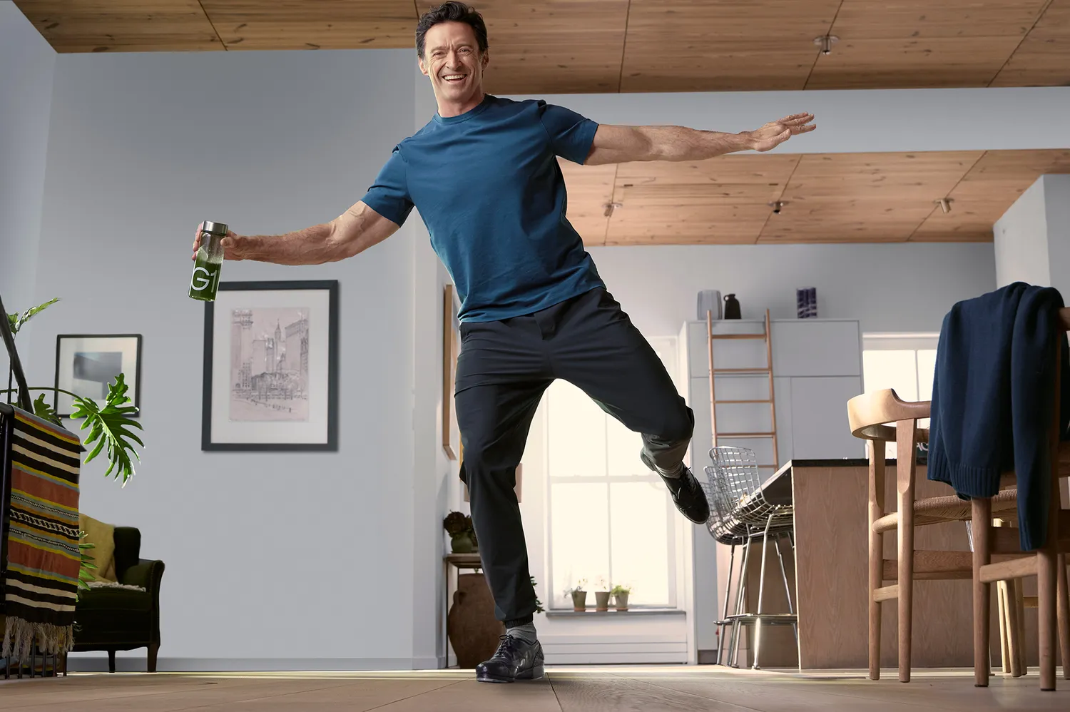 Hugh Jackman balancing on one foot while holding a can of AG1 supplement.