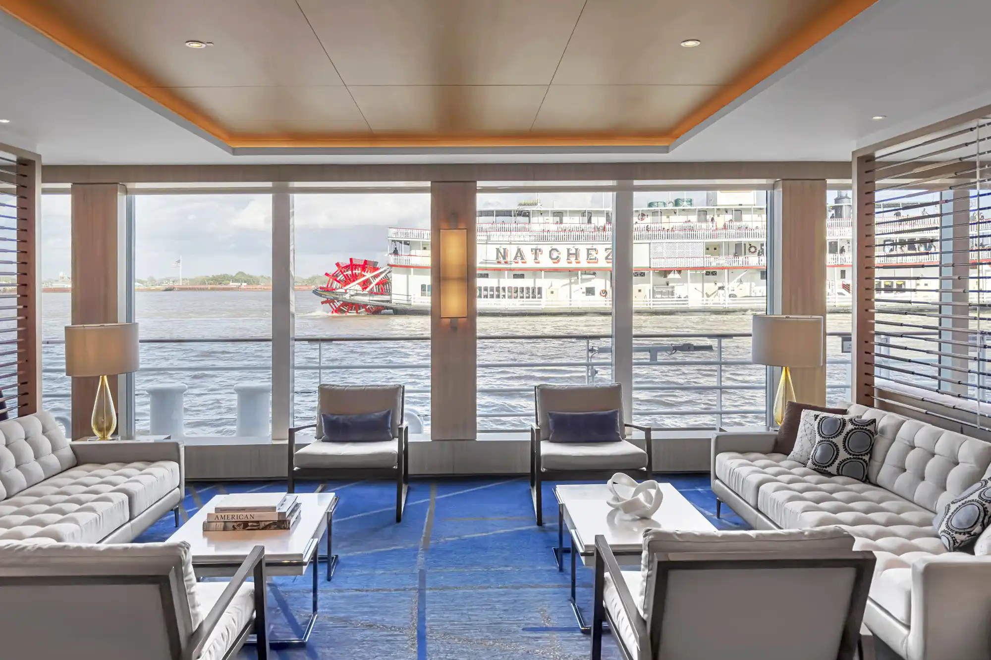 An interior lounge on a modern ship with large windows looking out at a traditional paddlewheel boat on the water.