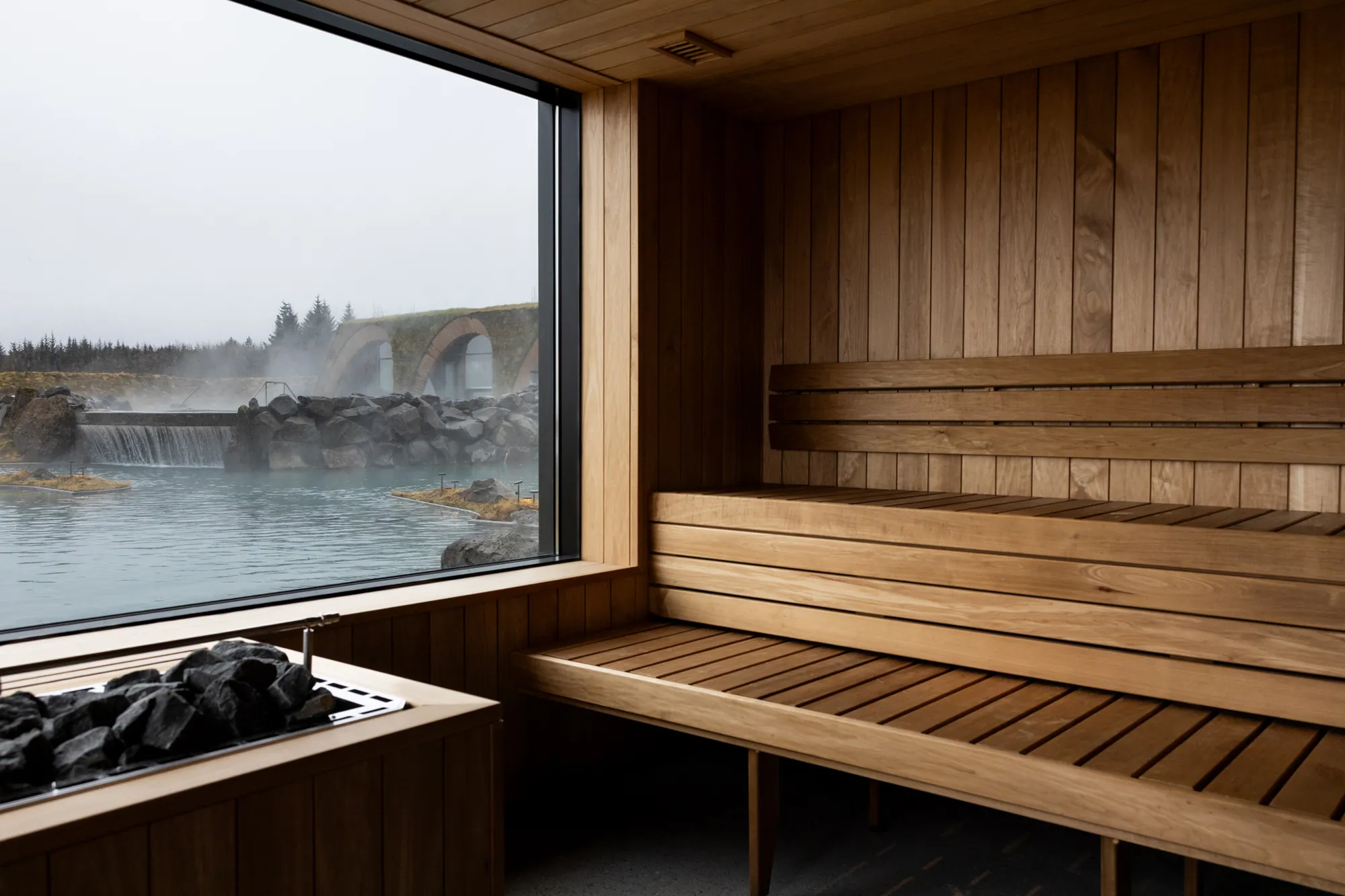 Interior of a modern wooden sauna with a large window overlooking the geothermal lagoon and waterfall.