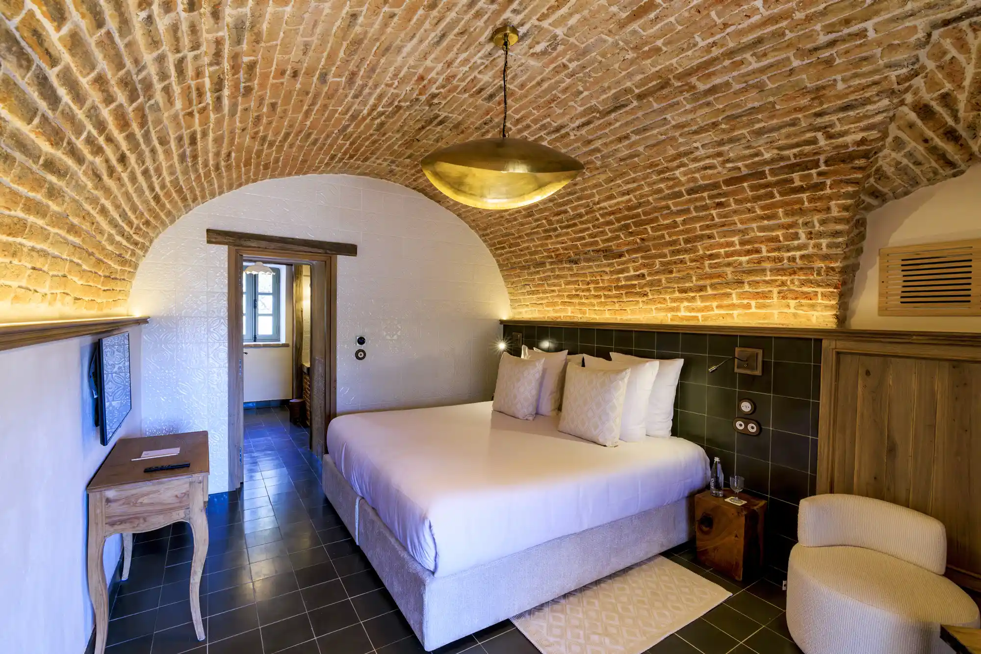 A luxury hotel bedroom with a vaulted brick ceiling and rustic-modern furniture in a converted barn setting.